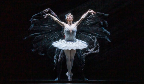 A dancer on stage wearing a white dress holds her arms in the air looking upwards The shadows of her arms moving up and down at the side of her