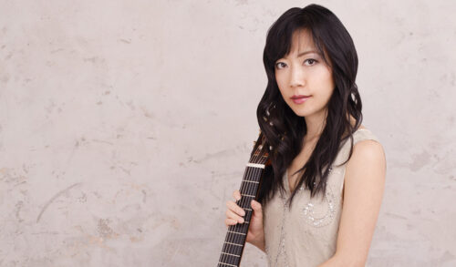 Image A woman stood in front of a concrete background holding the neck of a guitar