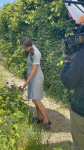 A girl in period dress standing looking at flowers. A man is filming her on a video camera.