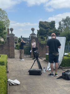Two people stand in front of a gate. A camera man is setting up a camera in front of them.