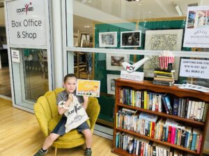 A boy sat on a chair in The Courtyard foyer holding a certificate and a tote bag next to a shelf of books