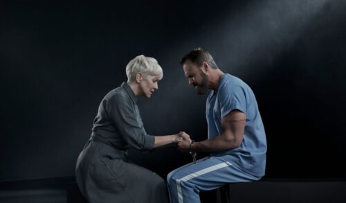 A woman with short white hair sits in front of a man in a blue prison uniform Their heads are bowed toward one another and they are holding hands The man has a pair of handcuffs on
