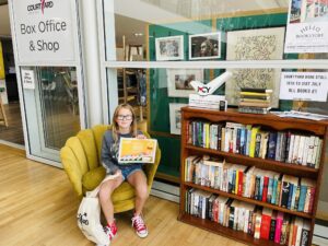 A girl sat on a chair in The Courtyard foyer holding a certificate and a tote bag next to a shelf of books
