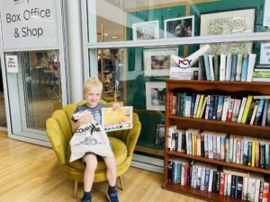 A young boy sitting on a chair next to a bookshelf. He is smiling and holding a certificate and a Courtyard tote bag.