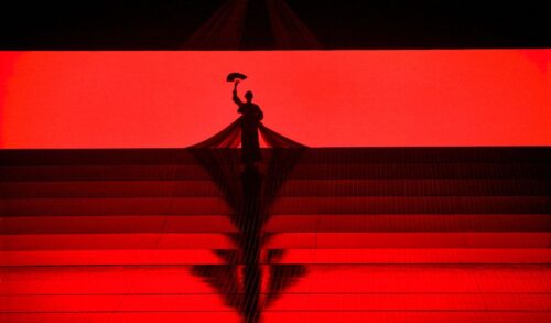 A stage is lit red A silhouette of a woman holding an umbrella appears at the very back of the stage
