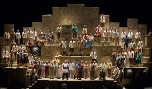 A large multitiered stage made up of light stone walls performers stand on each level facing the audience