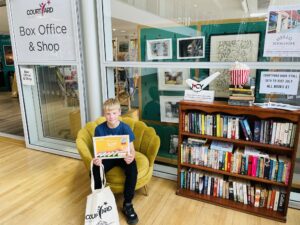 A boy sat on a chair in The Courtyard foyer holding a certificate and a tote bag next to a shelf of books