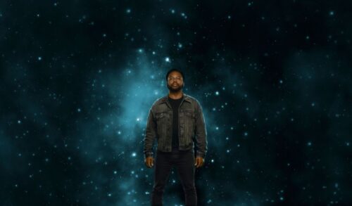 A man stands in a grey jacket Surrounding him is stars and a blue galaxy