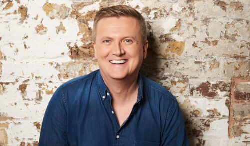 An image of Aled Jones standing against a brick wall smiling