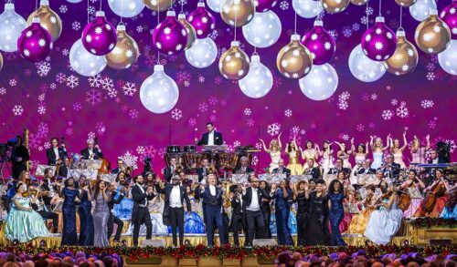A crowd of people stand on stage with Andre Rieu  there are Christmas baubles hanging from above