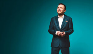 An image of Chris McCausland standing in a suit in front of a turquoise background