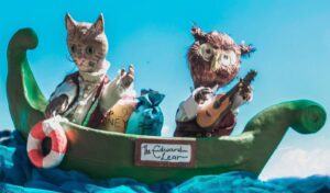 Puppets of an owl and a pussycat sit on a green boat. The boat is riding on blue waves made of fabric.