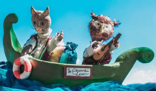 Puppets of an owl and a pussycat sit on a green boat The boat is riding on blue waves made of fabric