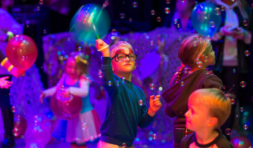 A young boy in glasses and wearing superhero face paint waves a balloon surrounded by bubbles