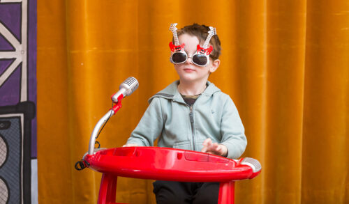 An image of a little boy playing a toy keyboard wearing guitar shaped sunglasses