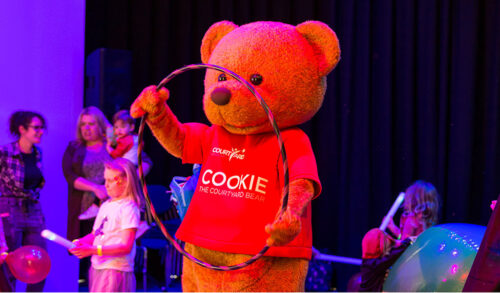 An image of Cookie the Courtyard bear holding a hula hoop under neon lights