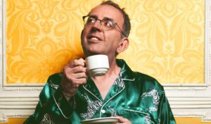 An image of Reverend Richard Coles wearing green satin pyjamas with zebras on. He is holding a cup and saucer and looking up smiling thoughtfully.
