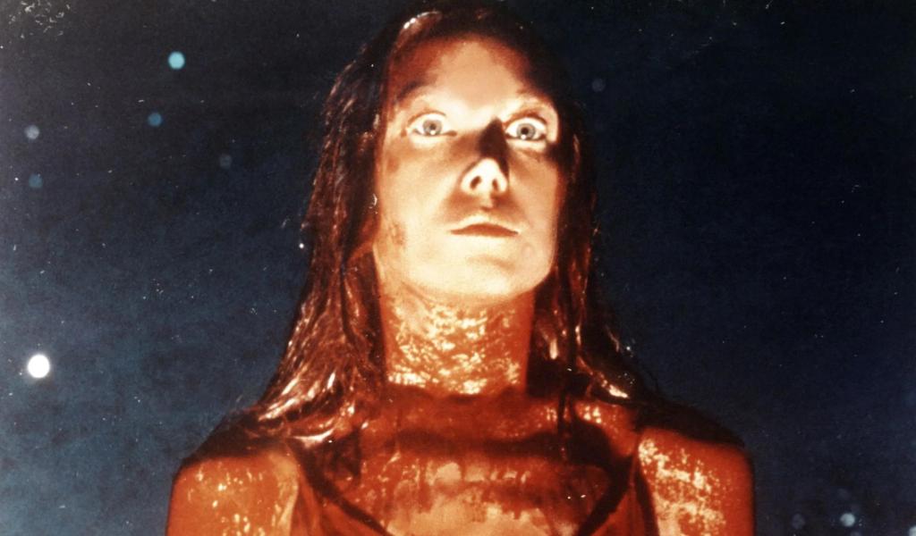 A close up image of a young woman in a prom dress, covered in blood. She is staring forward intensely. 