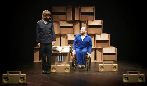 An image of two men wearing boiler suits one standing and one in a wheelchair in front of a stack of cardboard boxes