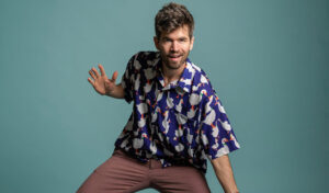 An image of a man in a patterned shirt holding his arms out and smiling to the camera against a teal background