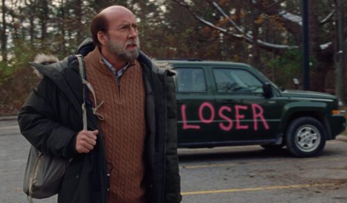 A man with a beard wearing a coat walks across a car park A car behind him has the word loser painted on the side of it