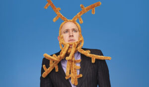 A man wearing a suit stands looking concerned. His head is wrapped up in reindeer headbands that look like antlers.