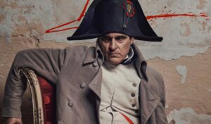 Joaquin Phoenix as Napoleon sits on a chair in his French military uniform. Behind him is a concrete wall with blood on it.
