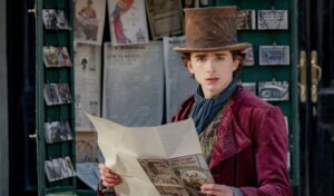 A young Willy Wonka stands in a purple coat and brown top hat outside a shop. He is holding a newspaper open in his hands.