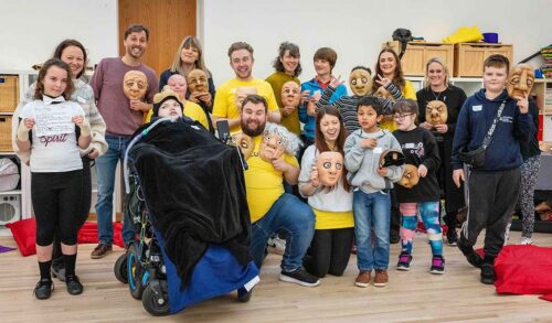 A group of disabled children pose and smile with The Courtyard team holding crafted masks of different faces