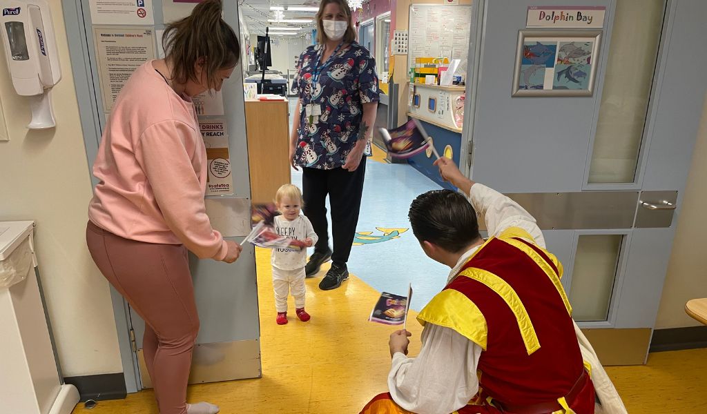 An actor dressed as Dick Whittington waving a flag opposite a very young girl, who is also waving a flag. Her mum and a nurse stand near them.