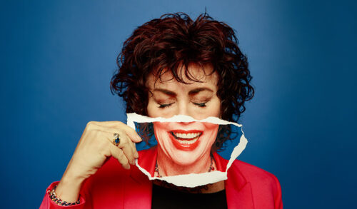An image of Ruby Wax a woman with dark curly hair holds up a piece of paper with an image of her mouth printed on it