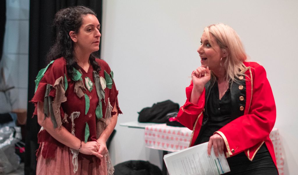 Rehearsal from Cinderella - a woman in a red jacket rests her chin on her hand and looks excitedly at a woman wearing a top covered in fabric leaves