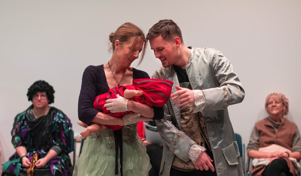 Two actors cradle an armful of clothes, resembling a baby. They look at it lovingly.