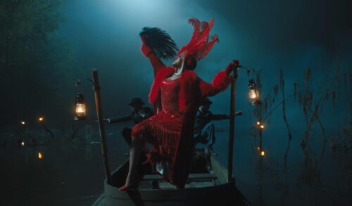 A woman dressed in red with a red feathered headdress poses on a dimly lit boat surrounded by lanterns and fog