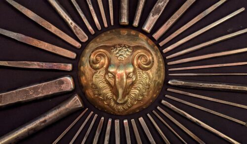 A close up image of a golden metal sun embedded in wood At the centre of the sun their is the head of a ram with curled horns
