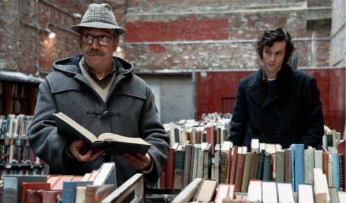 Two men stand surrounded by books One holds an open book and looks off into the distance He has a moustache wears glasses and has a tartan hat on The other stands behind looking at him