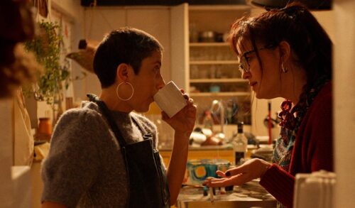 Two white people stand in a cluttered kitchen One has buzz cut hair and hoop earrings they are sipping from a white mug and making strong eye contact with the other who has glasses and dark red hair pulled into a ponytail The exchange seems serious