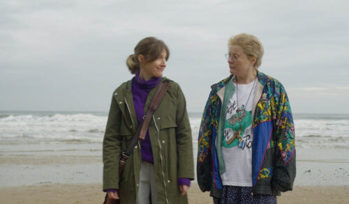 Two white people stand next to each other on a beach the weather is gloomy A white woman on the left is in a rain mac with her brown hair pulled into a ponytail She looks to the person next to her who is wearing a mismatched outfit with an 80s jogging jacket graphic print T shirt and spotty bottoms