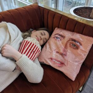 A woman lays on a sofa with a cuddly popcorn bucket and a sequin cushion with the face of Nicolas Cage on it. 