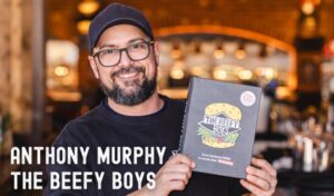 A man in a black t shirt and black cap stands smiling whilst holding a cook book.