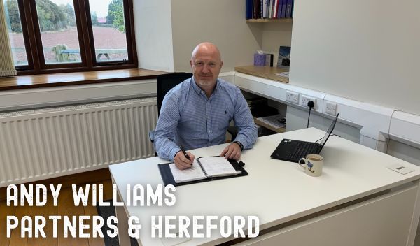 An image of a man sat writing at a desk, smiling at the camera. Writing reads 'Andy Williams Partners & Hereford'