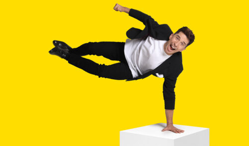 An image of comedian Russell Kane holding himself up on one arm on a white box