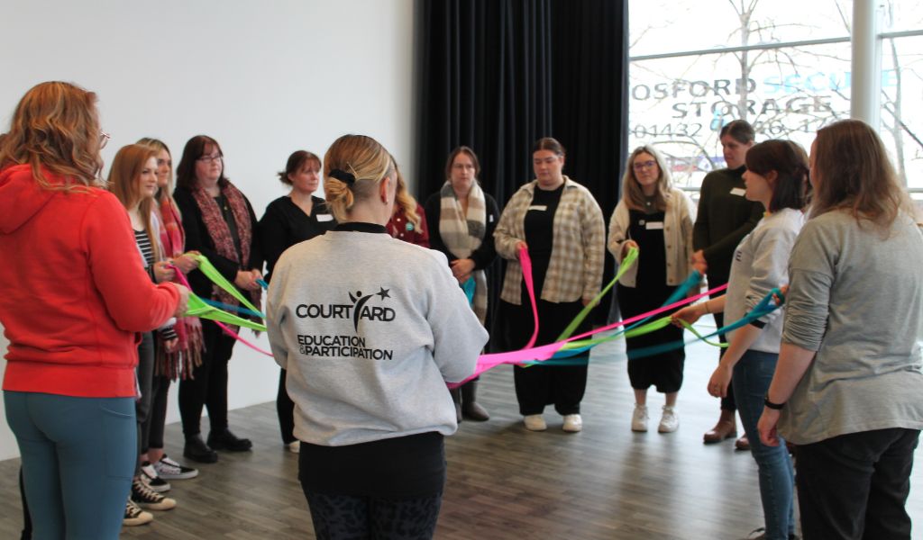 An image of a group of adults standing in a circle. They each hold a coloured ribbon.