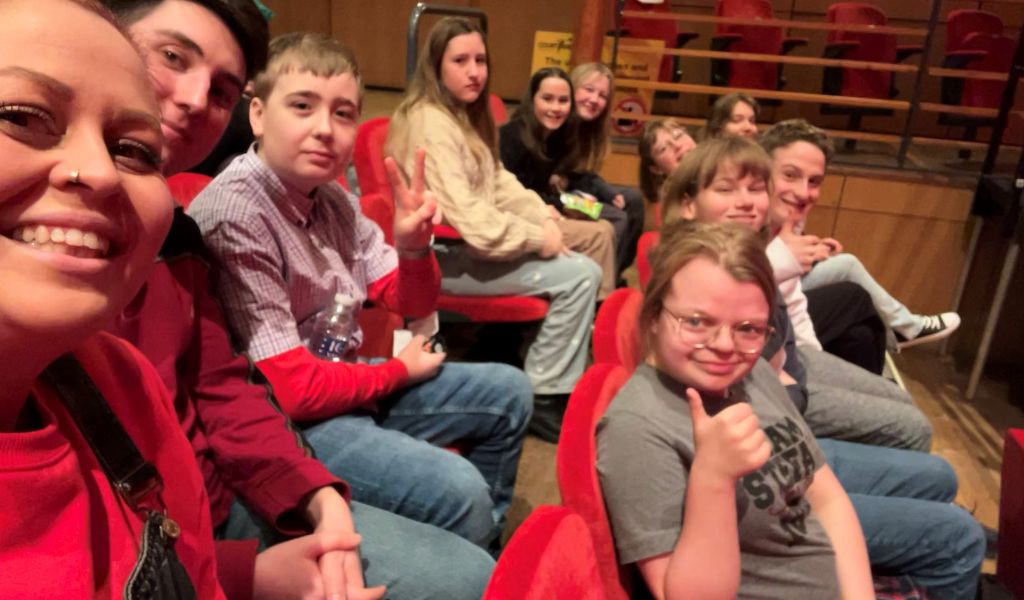 A selfie of young people and two Youth Theatre leaders sitting in an auditorium smiling