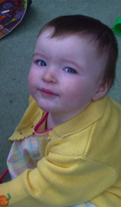 A very young girl wearing a yellow cardigan and smiling at the camera