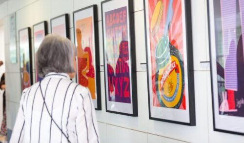 A woman stands and looks at a framed print In the print is a depiction of a marmite jar with the words bread sauce written on it