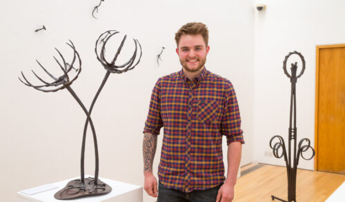 Artist Bran Davies stands in front of his artwork Behind him are a collection of metal sculptures which each represent a form of plant life