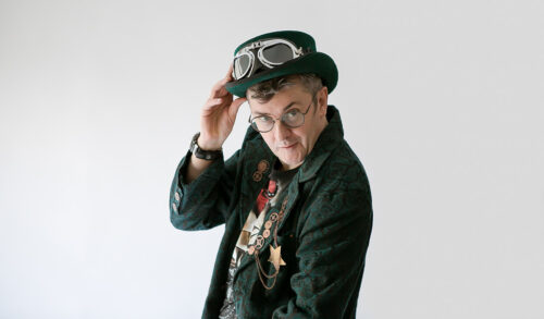 An image of Joe Pasquale tipping his hat with goggles on it and looking straight at the camera