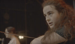A woman with curly hair has a determined expression on her face, she is preparing to strike with a sword.