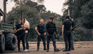 The film set of a military scene. Camera men are holding equipment off to one side while a group of men and one woman stand waiting for the next shot in their dark clothes and camo gear.
